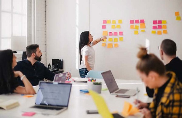 A professional woman stands at the front of a conference room pointing to a wall of sticky notes while her coworkers look on from a large table