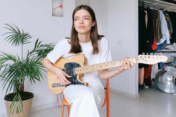 A young woman looks off into the distance while sitting in a minimalistic room and playing her guitar