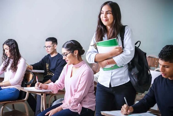 A female student standing with her books and bag among her peers in a classroom sitting down studying